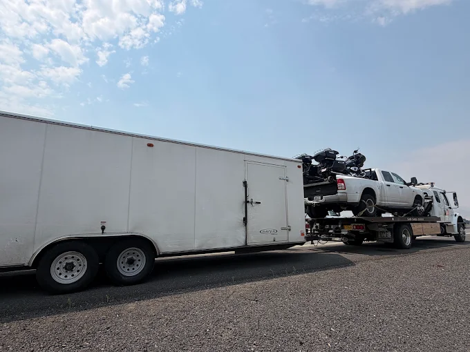 A flatbed with a loaded pickup on the deck, towing a large enclosed trailer on an open highway.
