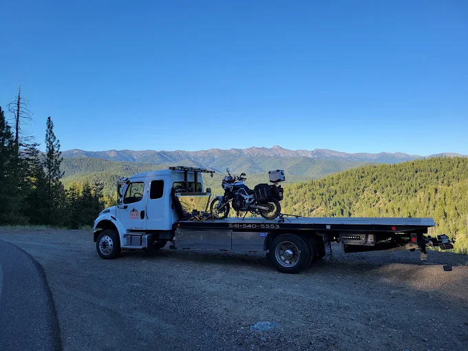 A flatbed on a mountain road carrying a loaded adventure motorcycle, with a forested mountain range behind.