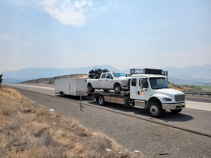 A flatbed carrying a pickup loaded with motorcycles, while towing an enclosed cargo trailer.