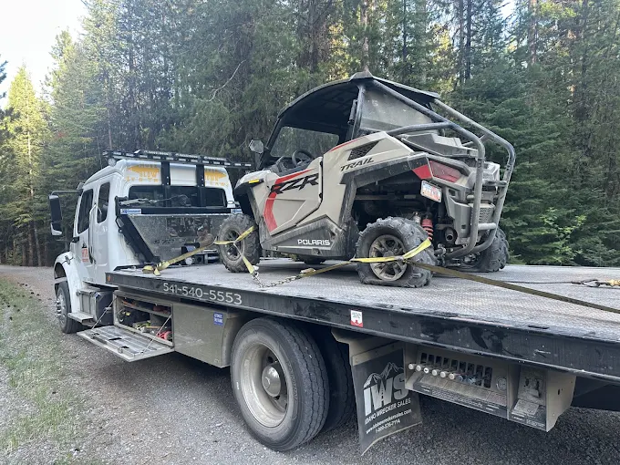 A flatbed in a forest carrying a damaged Polaris side-by-side, strapped down.
