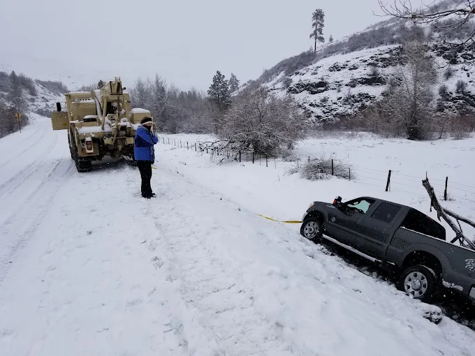 A heavy 6x6 recovery truck winching a pickup out of a snow-filled ditch during a snowstorm.